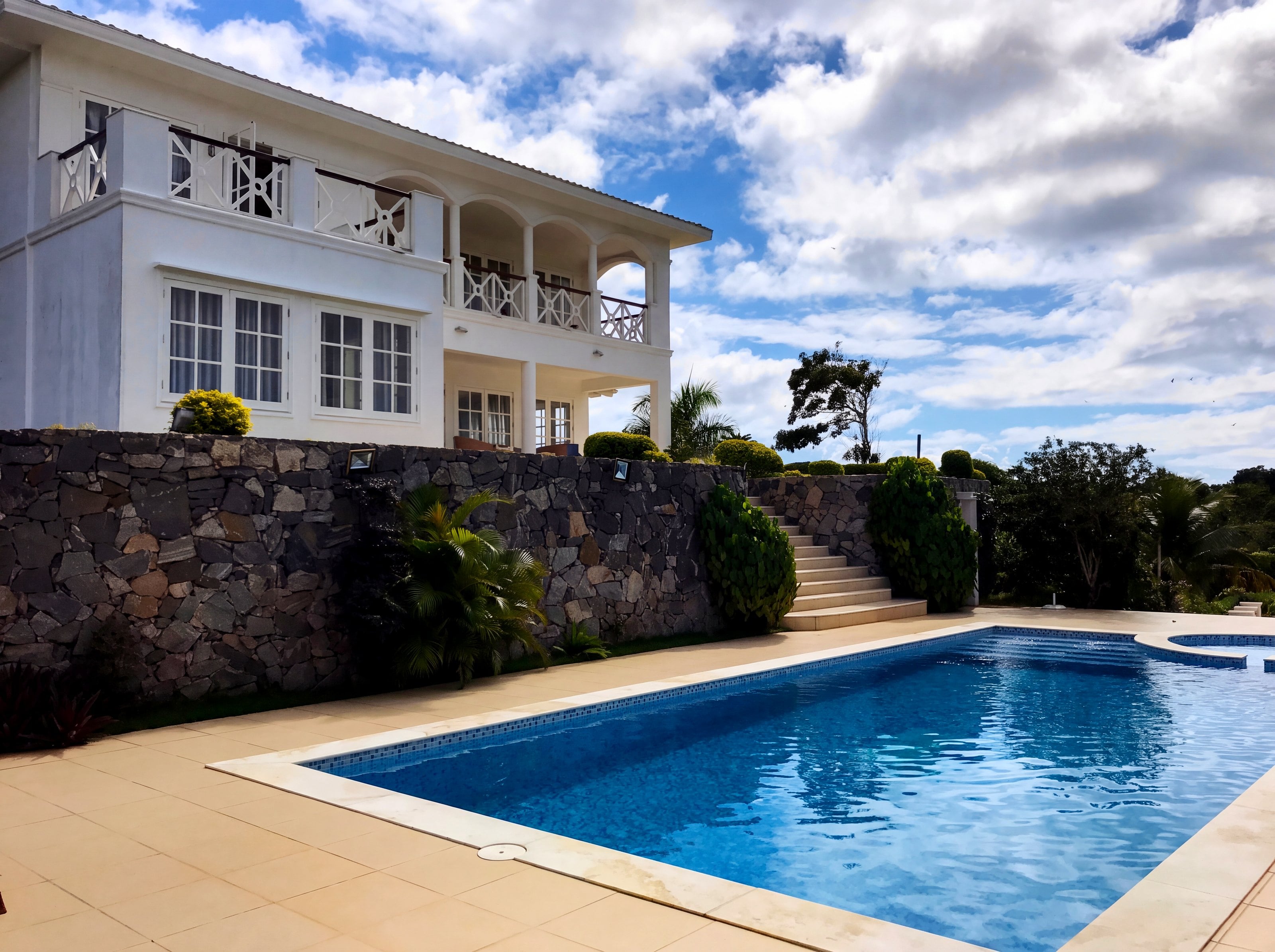 Casa branca de dois pavimentos com varanda em arcos, muro de pedra, escada e piscina azul sob céu com nuvens