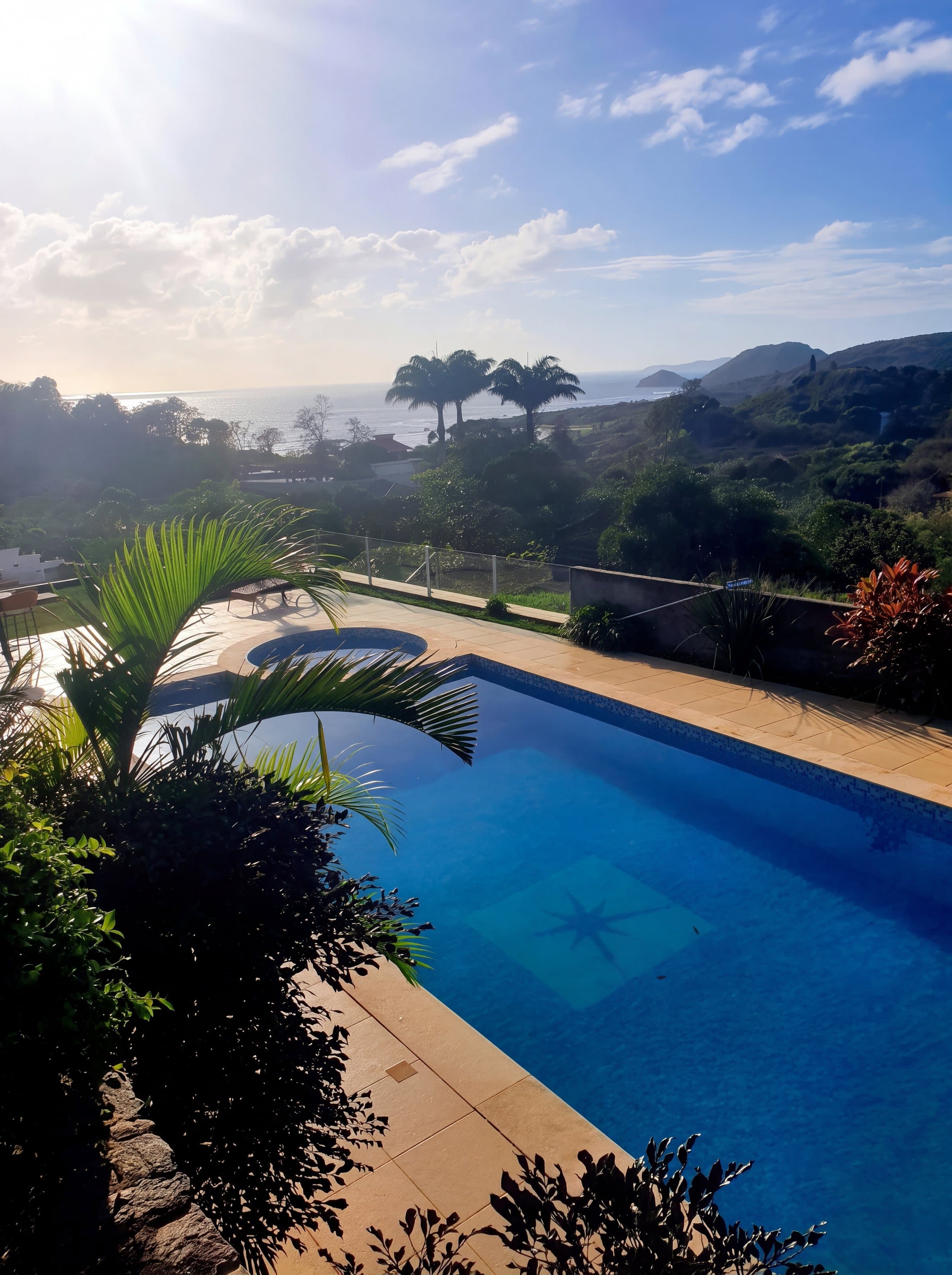 Piscina vista do alto, emoldurada por uma palmeira, com jardim e paisagem ao fundo ao entardecer